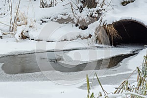 Winter landscape of a stream flowing out of a pipe, a frozen river With grass reeds Covered With Frost And Snow.