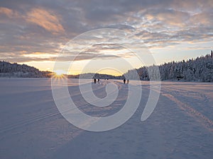 Winter landscape, sunset over a snowy forest and a frozen lake. Skiers and people walking in the background.