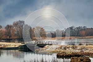 Winter landscape with trees near the river and cloudy sky with dim sun, bridge over the river. The beginning of winter, the first