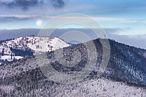 Winter landscape - view of the snowy forested mountains in the evening under the moon