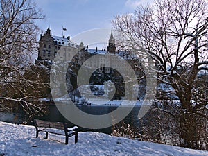 Winter landscape with wooden bench and bare trees on snow-covered shore of Danube River and Hohenzollern Castle in Sigmaringen.