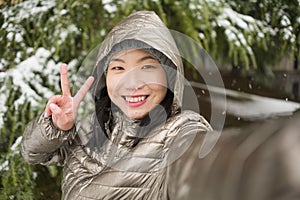 Winter lifestyle portrait of young happy and beautiful Asian Japanese woman taking selfie picture with mobile phone enjoying snow
