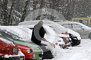 Winter. A man with a broom cleans car from snow on the street after big snowstorm in the city, all cars under snow, icy roads, sno