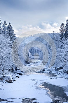 Winter mountains with Christmas trees strewn with snow, ice-bound river and clouds in the blue sky.