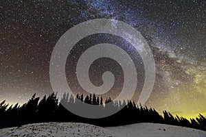 Winter mountains night landscape panorama. Milky Way bright constellation in dark blue starry sky over dark spruce pine trees