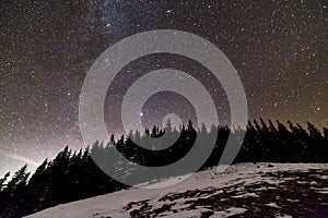 Winter mountains night landscape panorama. Milky Way bright constellation in dark blue starry sky over dark spruce pine trees