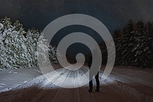 Winter night scene, a man with a flashlight in a snowy forest on the road, starry sky with clouds