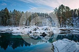 Winter panorama, small turquoise lake in the mountains among snow-covered forest. Trees are reflected in the lake water. Majestic