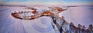 Winter panorama. Snow covered fields, meadows. River, frozen trees, village in evening light. Rural sunset landscape. Dirt road in