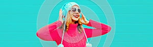 Winter portrait cheerful young woman in wireless headphones listening to music wearing pink knitted sweater and white hat with pom