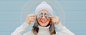 Winter portrait close up of happy smiling woman holding snowflakes covering her eyes wearing a white knitted hat over blue