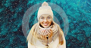 Winter portrait of happy smiling blonde young woman warms up holding mug of hot coffee or tea wearing hat, knitted sweater and