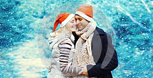 Winter portrait of happy smiling young couple hugging in red santa hats outdoors on christmas tree background with snowflakes