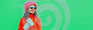 Winter portrait of happy smiling young woman looking away wearing knitted sweater, pink hat on green background