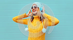 Winter portrait happy smiling young woman in wireless headphones listening to music wearing yellow knitted sweater and white hat