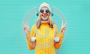 Winter portrait happy smiling young woman in wireless headphones listening to music wearing yellow knitted sweater and white hat