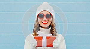 Winter portrait of happy young woman holding red gift box in her hands wearing a white knitted hat, heart shaped sunglasses