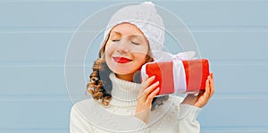 Winter portrait of happy young woman holding red gift box in her hands wearing a white knitted hat, sweater over blue
