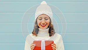 Winter portrait of happy young woman holding red gift box in her hands wearing a white knitted hat, sweater over blue