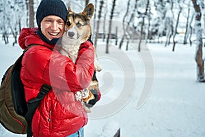 Winter portrait man wearing warm clothes embracing his pet Corgi dog in winter at snow park