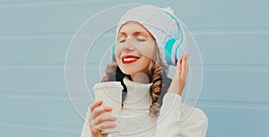 Winter portrait of smiling woman listening to music in headphones with coffee cup wearing a white hat, knitted sweater over blue