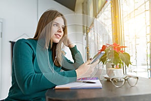 Winter portrait of student girl studying in cafe sitting with cup of coffee. Young woman studying using smartphone, listening to