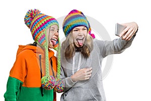 Winter portrait of two happy beautiful teenage girlfriends in knitted hats having fun with mobile phone, taking selfie, isolated o