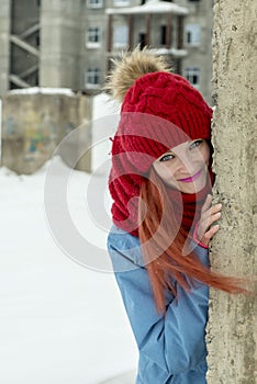 Winter portrait of young beautiful woman in red hat and red comforter hiding behind grunge concrete wall