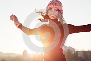 Winter portrait of a young smiling woman in a pink hat and mittens on a background sunset