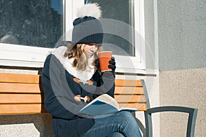 Winter portrait of young teen girl student in hat, warm clothes with cup of hot drink and book