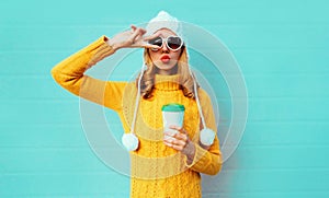 Winter portrait young woman with coffee cup showing peace gesture wearing yellow knitted sweater, white hat with pom pom