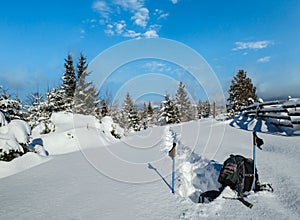 Winter remote alpine village outskirts, snow drifts on mountain fir forest edge. Tourist backpack on a freshly trodden hiking