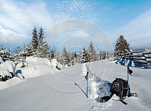Winter remote alpine village outskirts, snow drifts on mountain fir forest edge. Tourist backpack on a freshly trodden hiking
