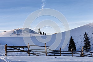 Winter rural background with small wooden alpine house, pines, fence, snow field, mountains, blue sky