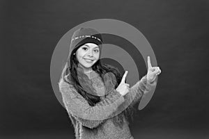 Winter sale. Happy girl in warm hat pointing index fingers at something. Small child smiling and pointing red background