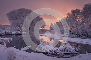 Winter scene at sunrise. Wintry morning with colorful sky on river. Beautiful snowy nature landscape. Amazing winter scenery with