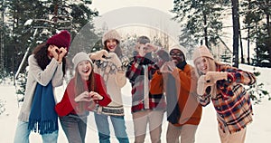 Winter season mood. Group of diverse happy friends look at camera smiling together, laughing at snowy forest slow motion