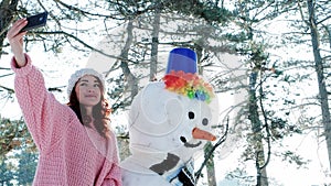 Winter selfie, cute girl making photo with snowman, a mobile phone in the hand of young woman making fun selfie photo in