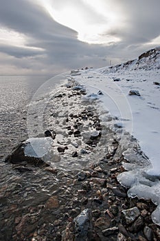 Winter shore of Lake Baikal with ice at the water`s edge and ice on stones in the water.