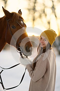 Winter side portrait of young smiling woman and brown horse. Woman with long hear in yellow cap holding snaffle of horse