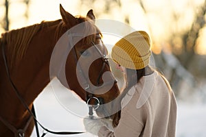 Winter side portrait of young woman and brown horse. Woman with long hear in yellow cap holding snaffle of horse
