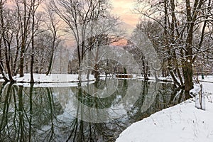Winter snowy landscape. A brick bridge leading over a calm river, which is lined with tall snow-covered trees reflecting on the