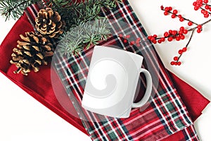 Winter still life. Blank coffee mug, fir tree branch, holly berries and pine cones. Checkered tartan plaid. Christmas