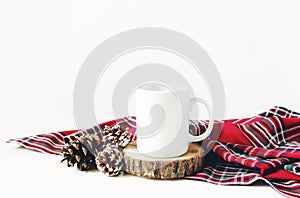 Winter still life composition. Blank ceramic coffee mugstanding on wooden cut board, pine cones and red checkered tartan