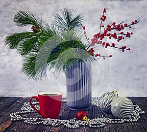 Winter still life with pine and ilex twigs in a vase on a light background and a red cup of coffee.