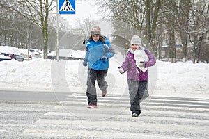 In winter, on the street in snowy weather, a boy and a girl cross the road along a pedestrian crossing