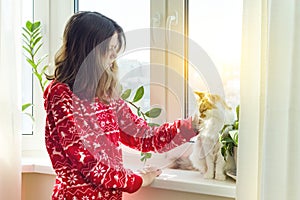 Winter time at home, young girl in winter warm pajamas with a cat looking out the window