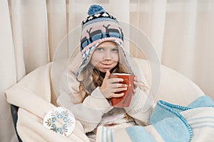 Winter time, little girl at home in an armchair with big cup of hot tea, in warm knitted hat under woolen blanket