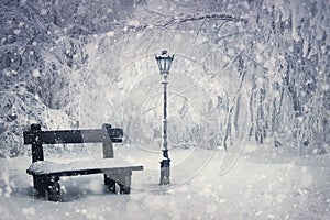 Winter time scene with a wooden bench and street lamp covered with snow under a snowfall in the park. Wonderful holiday season