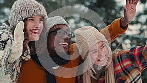 Winter vacation. Close-up three happy multiethnic young friends waving, smiling cheerfully at snowy forest slow motion.